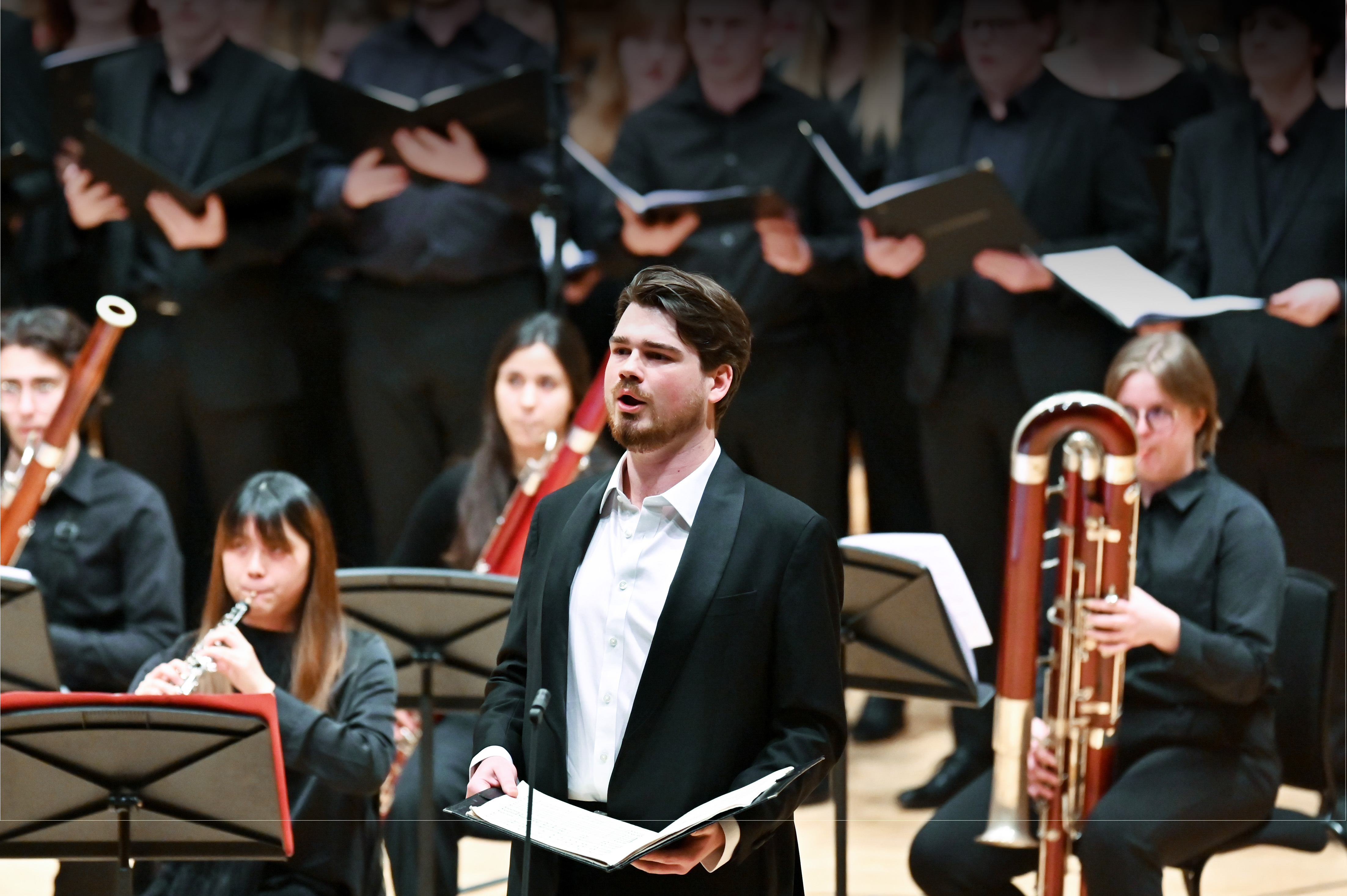 Male student, dressed in a formal attire, performing in an orchestra, with musicians and the choir in the background,